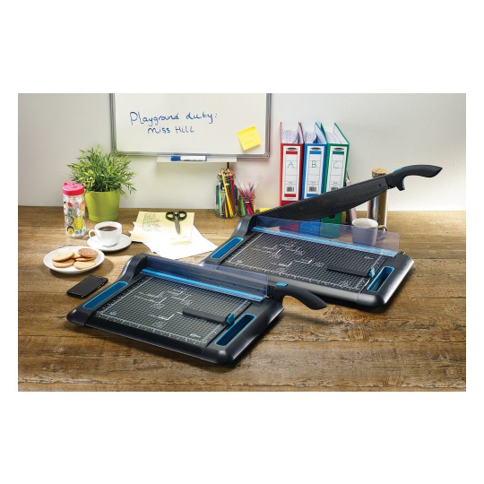 The image shows a desk with two paper cutters, a plant pot, a cup of coffee, cookies, a mobile phone, scissors, colored pens, and folders. There is also a whiteboard with a message.