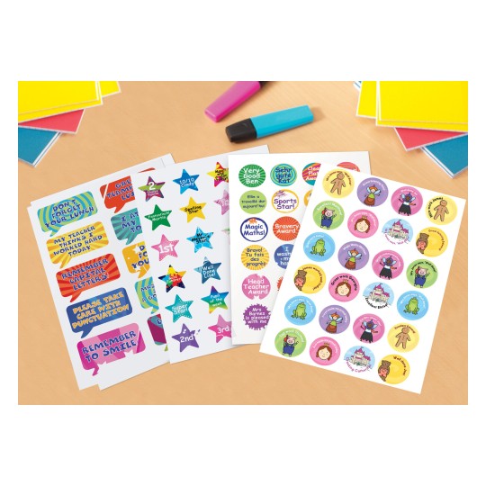 The image features various reward stickers with colorful designs and messages, such as 'Well Done', 'Sports Star', and 'Excellent Work'. They are laid out on a table alongside colorful paper and markers.