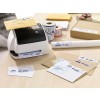 The image shows a label printer with envelopes and packages on a desk. There are various mail items, including yellow and white envelopes, packing tape, and rolls of labels.