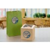 The image features a green paper bag and a brown cardboard box, both branded with 'Radiant Planet Café.' The bag has handles, while the box appears to contain a baked item. The background suggests a café setting with tables and plants.