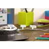 The image shows a laptop on a wooden desk with labeled cords for a phone charger, monitor, and printer. There are colorful pencils, a green stapler, and a green container with pens in the background.