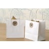 The image features two white gift bags with 'Thank You' labels on them. They are placed on a wooden surface, with a floral-patterned wall in the background. The bags contain items that are not fully visible.