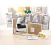 The image shows a packaging station with a printer, rolled paper, various envelopes, a cardboard box, and franking labels. In the background, there are bags, likely for shipping materials.