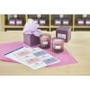 The image shows two scented candles placed in front of a purple gift box with tissue paper. There are also labels laid out on the table, indicating different candle scents. The background contains more purple boxes.