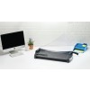 The image shows a workspace featuring an iMac computer, a plant in a pot, a white mug, a paper cutter, and a blue organizer with stationery items on a white table against a white brick background.