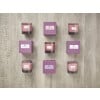The image features a collection of scented candles arranged in a grid pattern. Each candle is labeled with its fragrance name, such as 'White Lily', 'Cashmere', 'Pink Rose', and 'Lavender'. The candles are paired with matching purple boxes labeled 'Floral Nature'. The background is a light wooden surface.