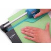 A person using a paper cutter designed by Avery to cut a green piece of paper. The cutter has a blue handle and a clear guide for precision.