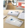 The image shows a stack of white envelopes with labels on them, placed on a desk with a keyboard and a notepad. A computer screen displays the Royal Mail website, indicating services for personal and business mail.