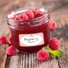 The image shows a jar of raspberry jam with fresh raspberries around it, resting on a wooden surface.