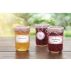The image features three jars of homemade jams placed on a wooden table. The jars contain marmalade, plum jam, and strawberry jam, each with a labeled tag and decorative twine. The background is softly blurred, suggesting a natural outdoor setting.