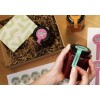 The image shows a gift box containing jars of raspberry jam and strawberry jam, with a thank you card and stickers. A hand is holding a jar that has a green label indicating it's raspberry.