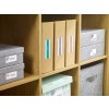 The image shows a wooden shelf containing various storage boxes and files. The boxes are labeled with words like 'Photos', 'Envelopes', 'Cards', and 'Expenses', indicating organized storage. The design includes patterns and colors, enhancing aesthetic appeal.