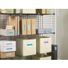 The image shows a wire shelving unit filled with organized storage boxes and file folders. The boxes are labeled with different categories such as 'Documents', 'Invoices', and 'Envelopes'. Some boxes are in neutral colors like beige and white, while others are in brown cardboard. The shelf serves as a functional storage solution for paperwork and supplies.
