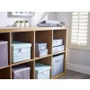 The image features a wooden shelf with various decorative storage boxes labeled for organization. Some boxes are designed in soft colors with patterns, and there are small plant decorations on the upper shelf. Sunlight is streaming through a window with louvered shutters.