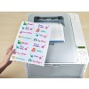 The image shows a hand holding a sheet of colorful labels printed with various names and designs, taken from a printer. The labels include items like 'Puzzles,' 'Ben's Ruler,' and 'Emma's Lunchbox' amongst stars and cartoon illustrations.