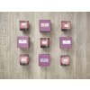 The image features decorative candles arranged on a wooden surface. There are four candles in glass holders labeled with scents such as 'White Lilac,' 'Lime & Basil,' 'Pink Rose,' and 'Lavender.' Each candle is paired with a matching purple packaging box, adding to the aesthetic appeal.