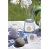 The image features a tranquil setting with a glass water pitcher, a white candle, a smooth stone, and a name card against a backdrop of a white tablecloth and green grass. Decorative symbols are placed on the stone and the card. White orchids are also visible, adding a serene touch to the scene.