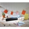 The image shows a creative workspace with various colored papers, pens, paper clips, a plant in a pot, and colored cards hanging on a string. There is also a device visible that is likely used for laminating.
