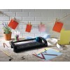 The image shows a creative workspace with a cutting machine, colored papers, pencils, washi tape, and a plant. Various colored cards are hanging on a string.