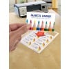 The image shows a printer and a hand holding a business card with the name 'Marcel Linder', while on the table there are labels with artistic styles. Colored pencils are a central element of the design.
