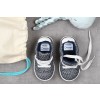 The image features a pair of small, gray knitted children's shoes with white laces, labeled 'TOM BRUNET.' Next to the shoes are a pastel-colored toy and a beige drawstring bag.