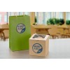 The image features a green paper bag and a brown cardboard box, both branded with 'Radiant Planet Café.' The bag has handles, while the box appears to contain a baked item. The background suggests a café setting with tables and plants.