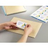 The image shows a hand applying a business card with a logo and address to a brown envelope. In the background, more envelopes and stickers are visible.