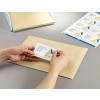 The image shows a person's hand applying a label to an envelope. The label contains a name and address, while a sheet of similar labels is visible in the background.