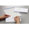 The image shows a person's hand applying a sticker label onto a white envelope. There is a sheet of label stickers with multiple designs visible, featuring a logo with a triangular shape and the text 'ABSTRACT'. Some envelopes are stacked in the background.