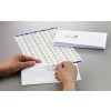 The image shows a person applying a label from a sheet of address labels onto a white envelope. The labels are arranged in a grid and have a logo with colorful geometric shapes. Several envelopes are stacked beside the sheet of labels on a gray surface.