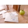 The image shows a wooden table with two addressed envelopes, a green apple, a glass container with wooden sticks, and a notebook. The envelopes have labels indicating names and addresses in France and Germany.