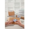 The image shows a desk with a few packaged shipments, including a box and an envelope. One hand is attaching a label to the envelope while another hand is involved. The background contains organized office supplies.