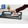 In the image, a worktable with a cutting tool is observed. A person is using a cutter to work with a sheet of pink paper on the surface. There is also a computer and a potted plant in the background.