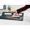 The image shows a hand using a cutting plotter or paper cutting machine. The device is on a table, with a purple piece of paper being cut, and in the background, there is a coffee mug and a plant.