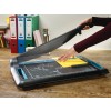 The image shows a hand using a paper cutter on a wooden table. In the background, there are colored folders and a pen holder.