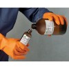 The image shows a person in orange gloves pouring a liquid from a larger brown bottle into a smaller brown bottle. The labels on both bottles contain warning signs and safety information.